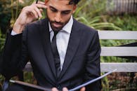 一名衣冠楚楚、身着黑色西装的男子，正聚精会神地坐在户外长凳上读书，散发着专业精神