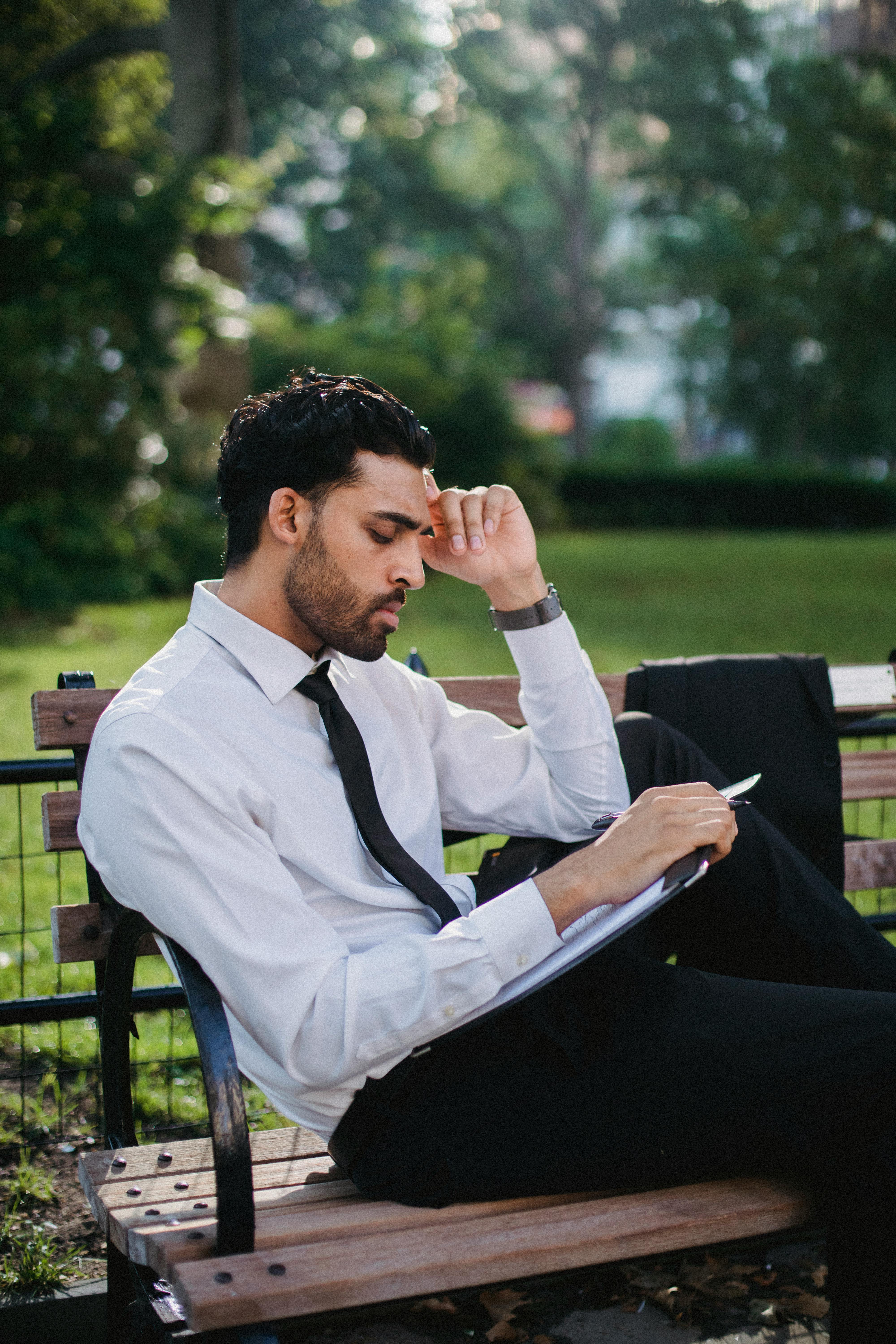 一名留着胡须、身穿正装的男子若有所思地坐在公园的木凳上读书