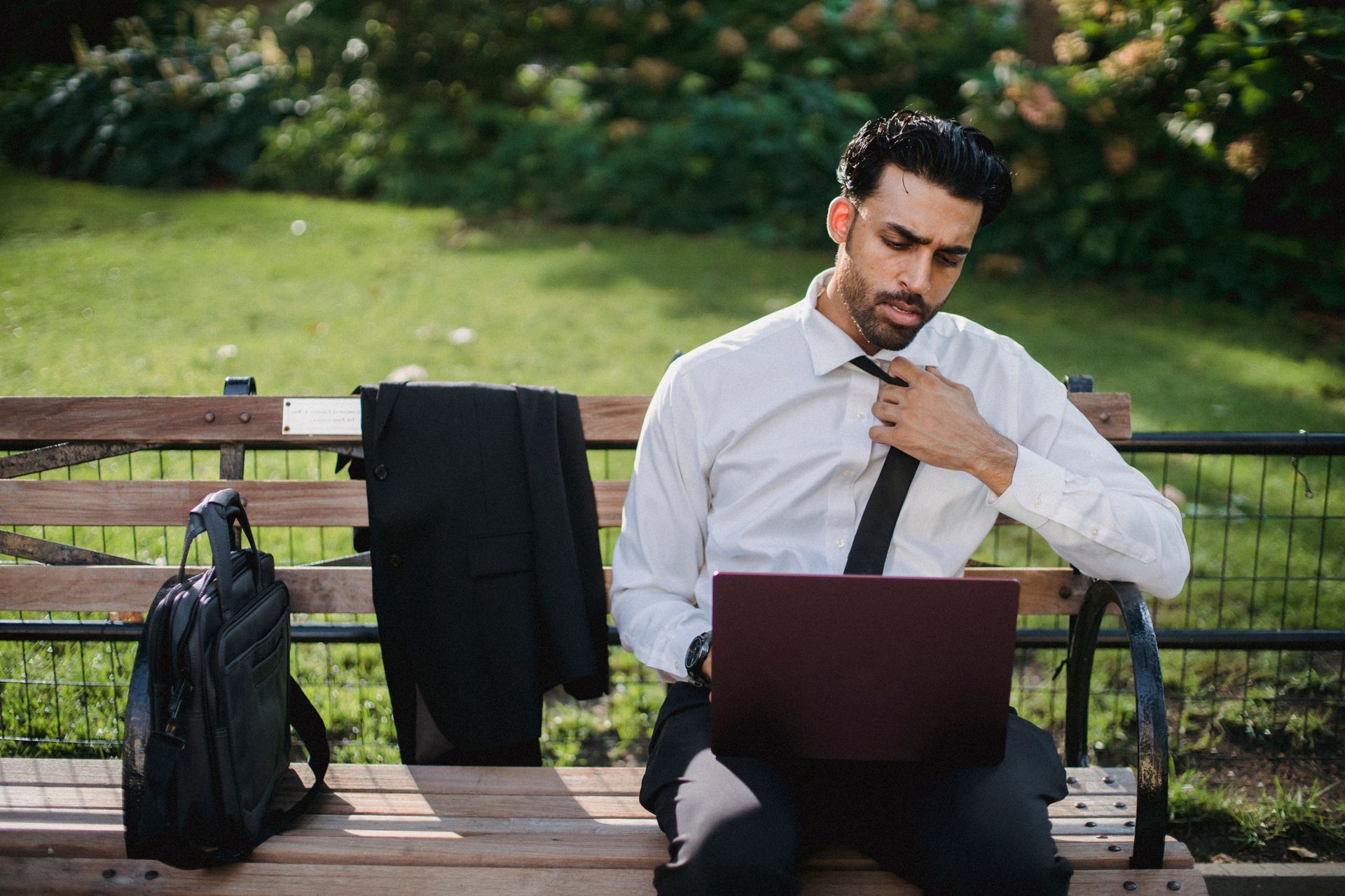 A businessman in formal attire working remotely on a laptop while seated on an outdoor bench.