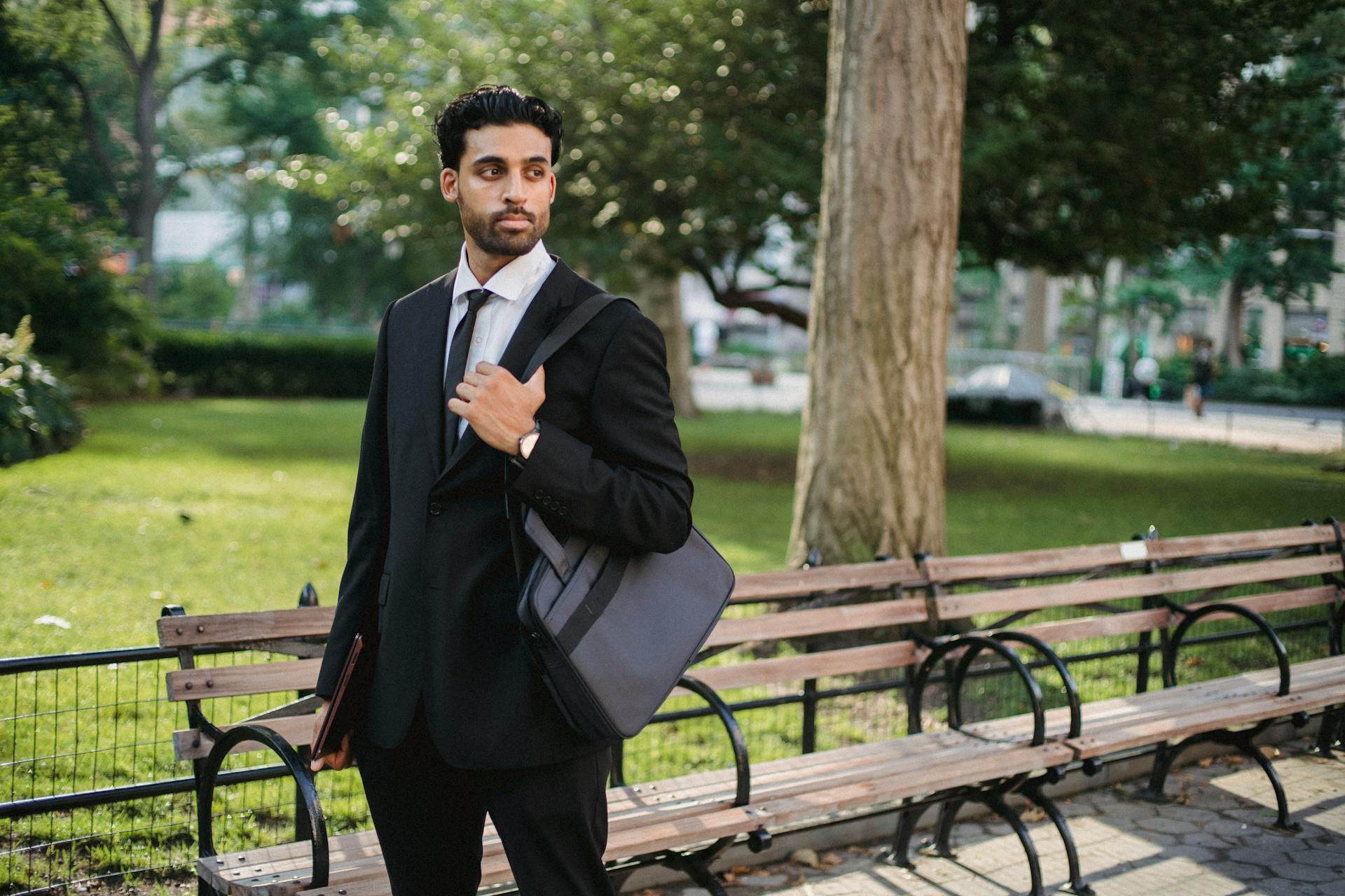 A business professional in a black suit holding a bag, standing in a park.