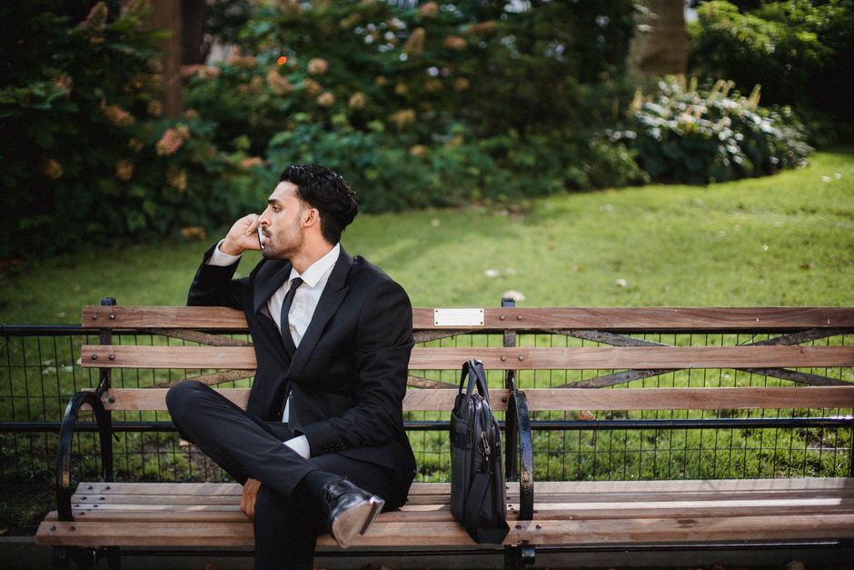 A well-dressed man in a black suit sits on a park bench talking on the phone.