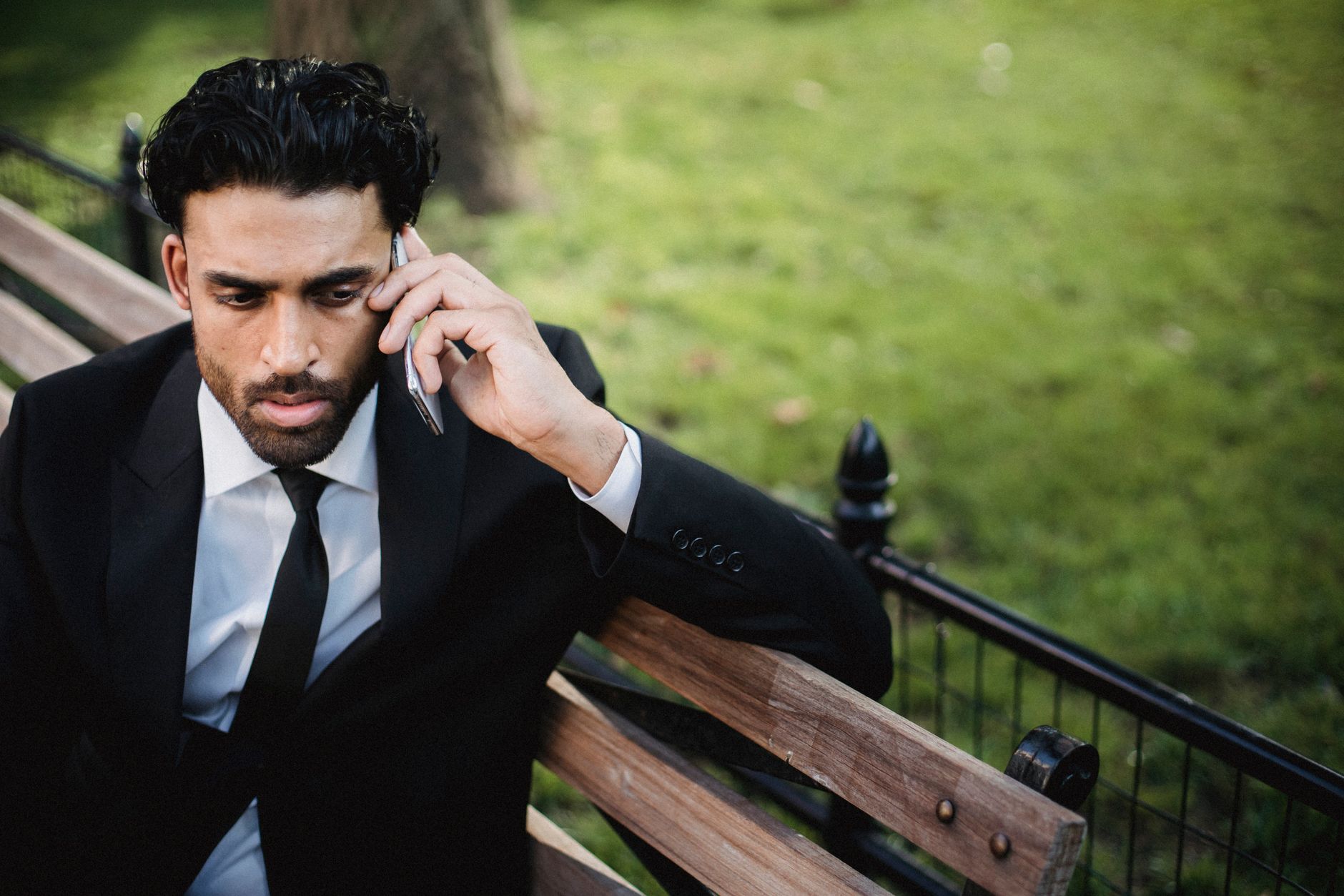 A man in a black suit sitting on a bench, engaged in a serious phone conversation in a park.