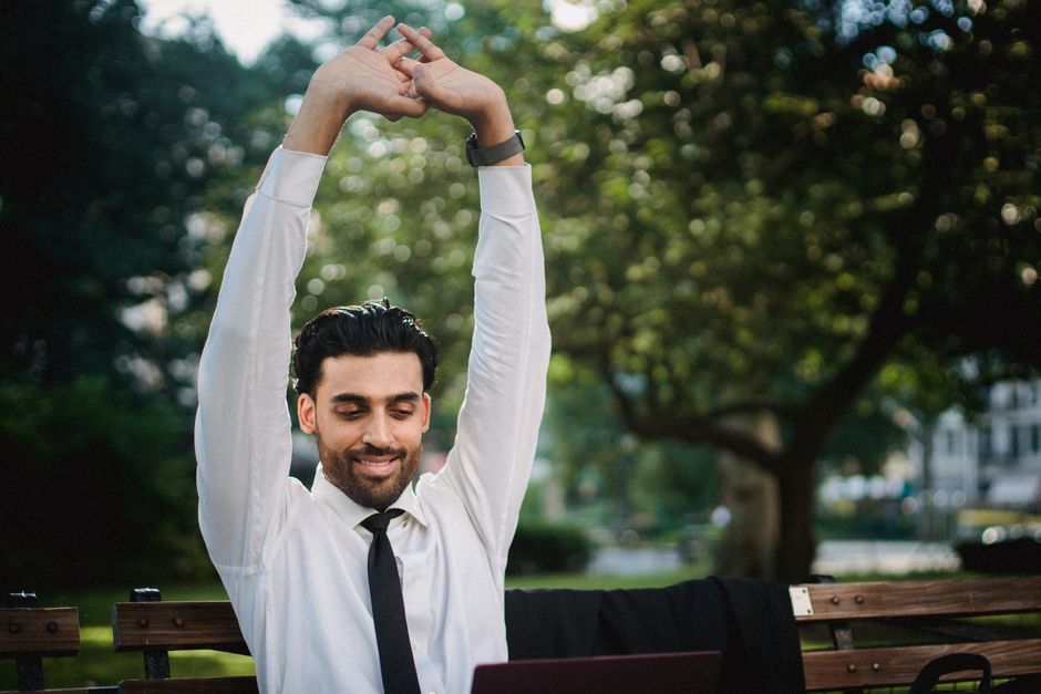 Smiling businessman stretches arms while sitting on park bench, wearing white shirt and tie.