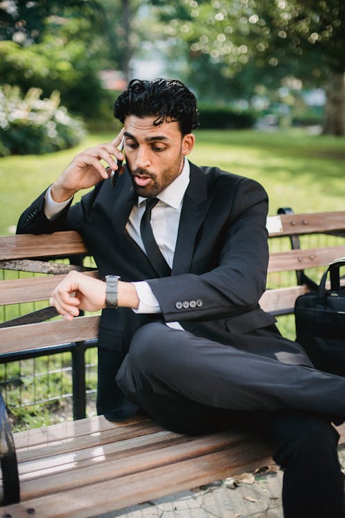 Man in Black Suit Sitting Bench Talking on the Phone and Looking His Watch