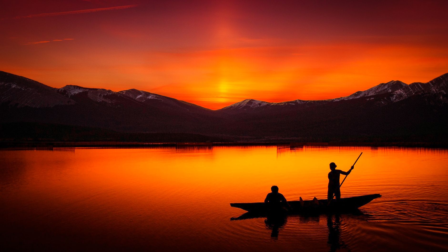 Silhouetted canoeists paddle under a vibrant sunset, casting reflections on a tranquil lake.