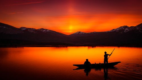 Free Silhouetted canoeists paddle under a vibrant sunset, casting reflections on a tranquil lake. Stock Photo