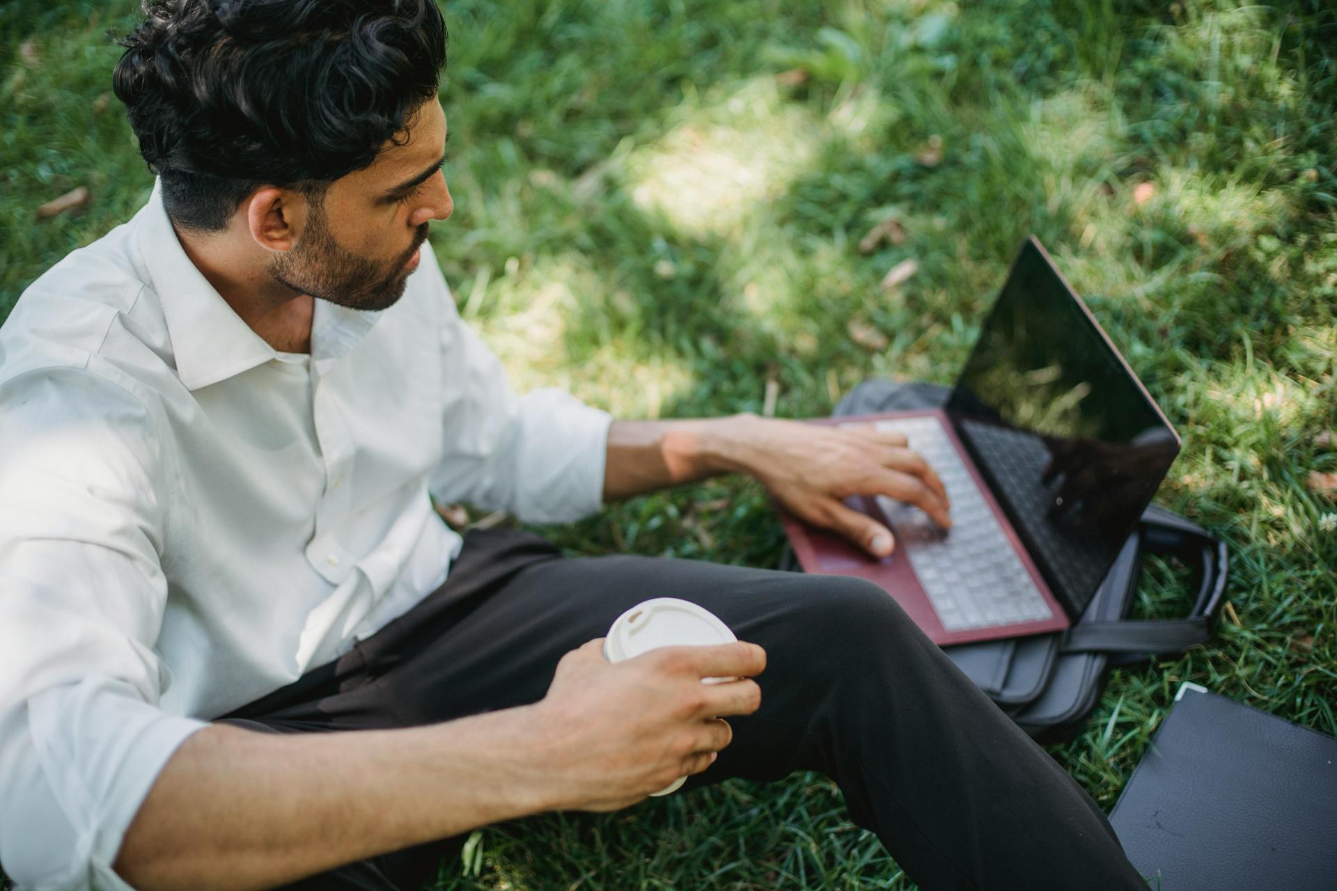 Man working on a laptop while sitting on grass, holding coffee. Perfect for remote work lifestyle visuals.