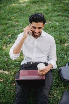Businessman working remotely outdoors with a laptop and phone, enjoying a coffee break.
