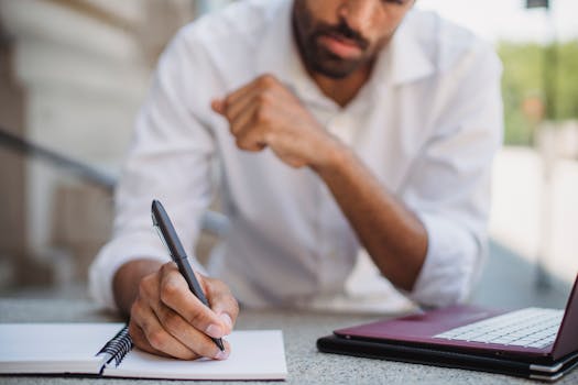 Man writing in notebook with a pen beside a laptop outdoors.