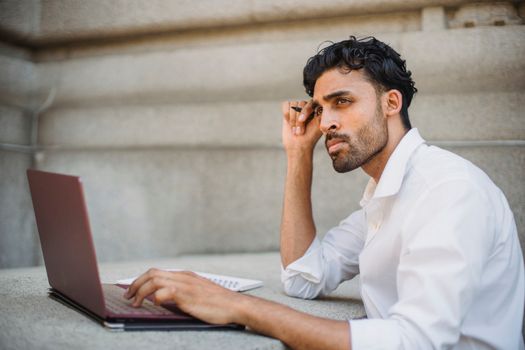 Professional man in a white shirt working on a laptop, contemplating ideas in an urban setting.
