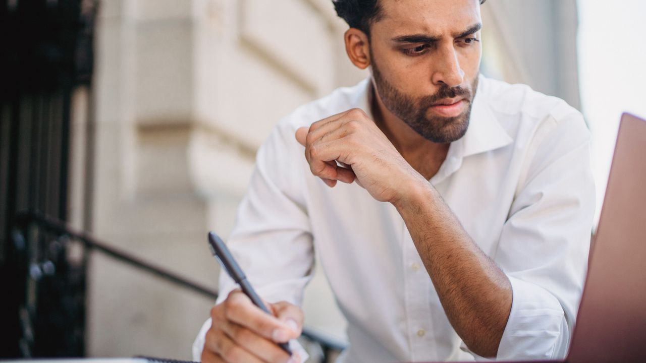 Focused man in white shirt takes notes while working remotely on his laptop outside, exuding concentration.