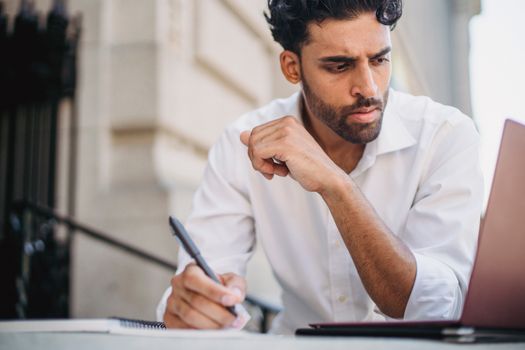 Focused man in white shirt takes notes while working remotely on his laptop outside, exuding concentration.