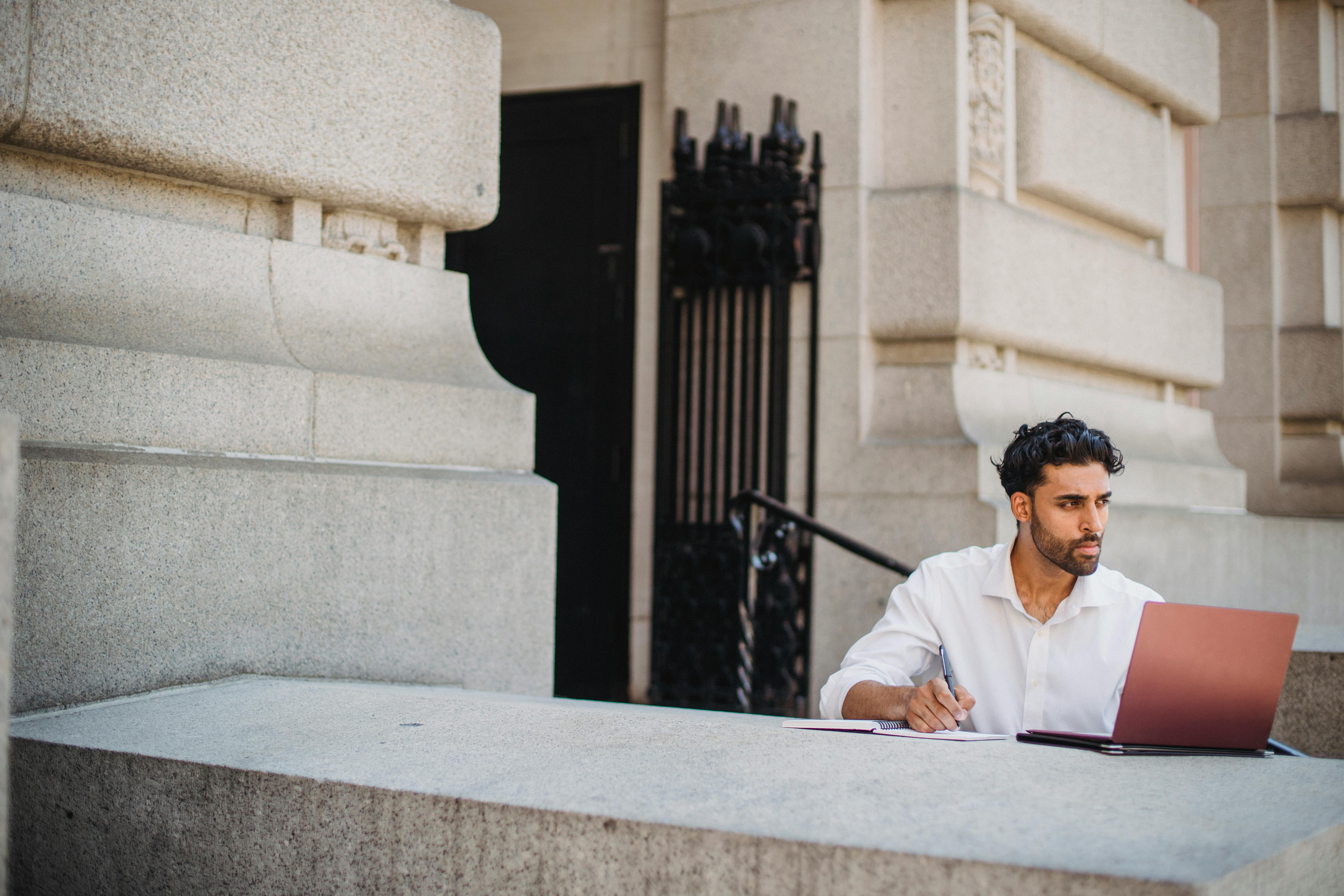 Businessman using a laptop and taking notes outside a stone building, symbolizing modernity and tradition.