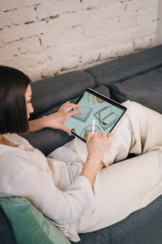A woman using a tablet and stylus for design work while relaxing on a couch in a cozy living room.