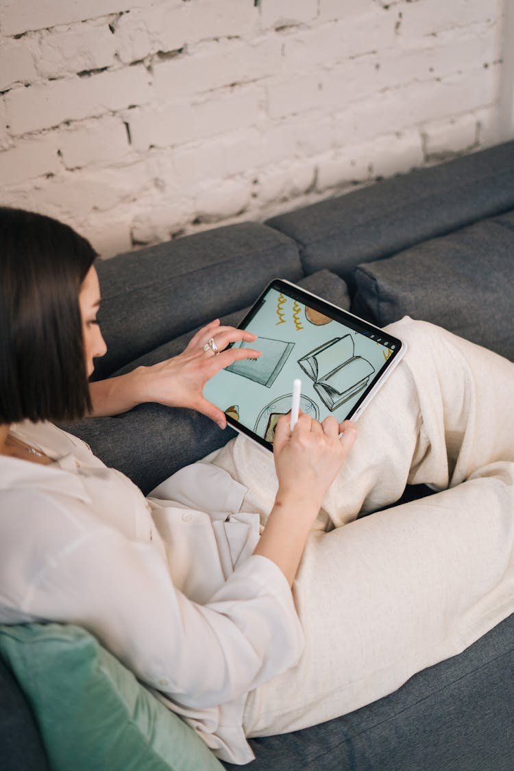 A Woman Sitting On The Couch While Drawing On The Ipad 