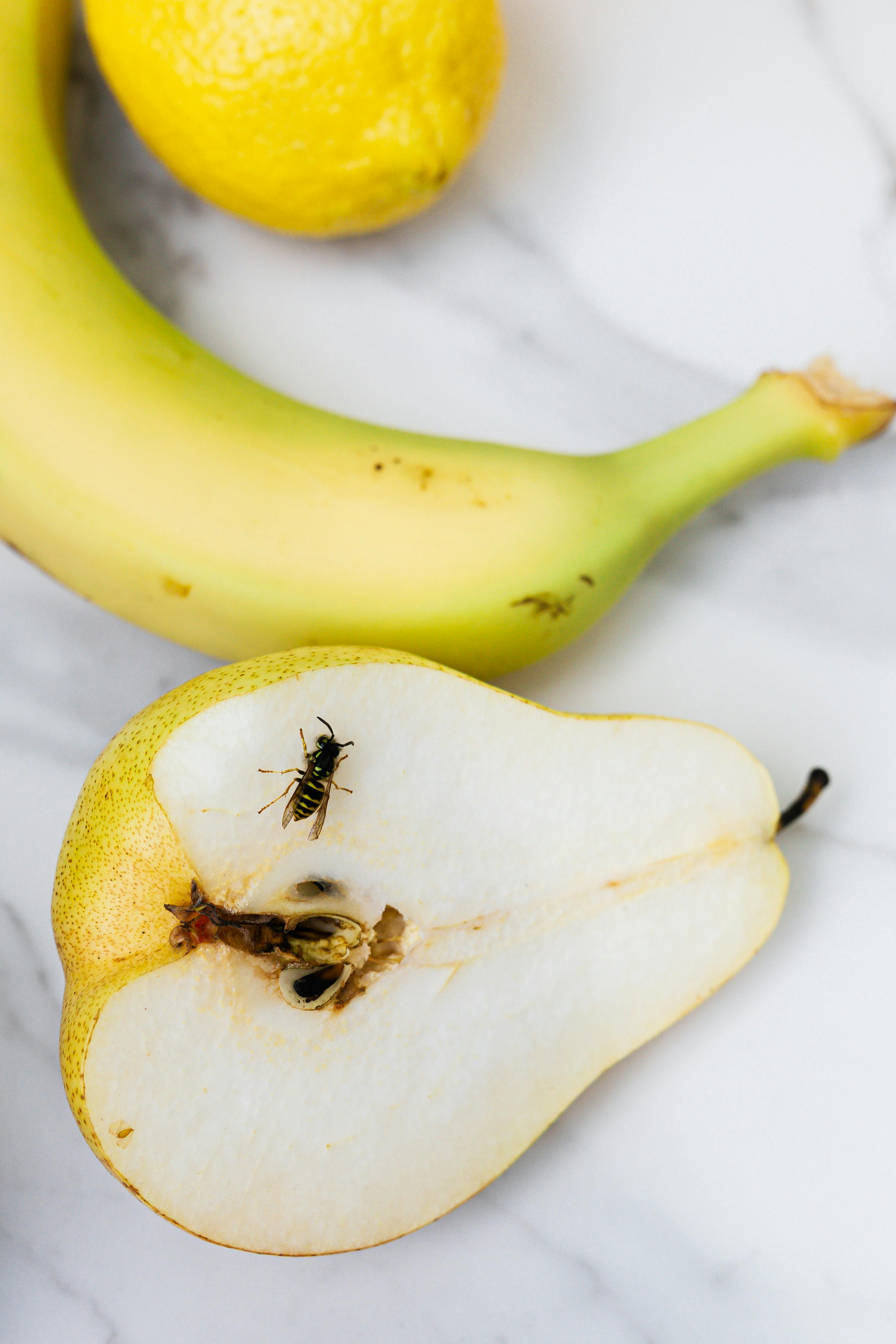 A wasp sits on a sliced pear next to a banana and lemon on a marble surface.