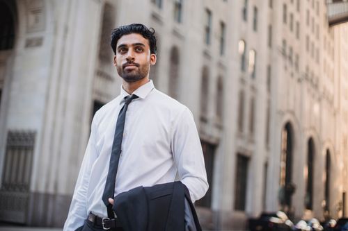 Free Young businessman with jacket over shoulder in cityscape background, looking confident. Stock Photo