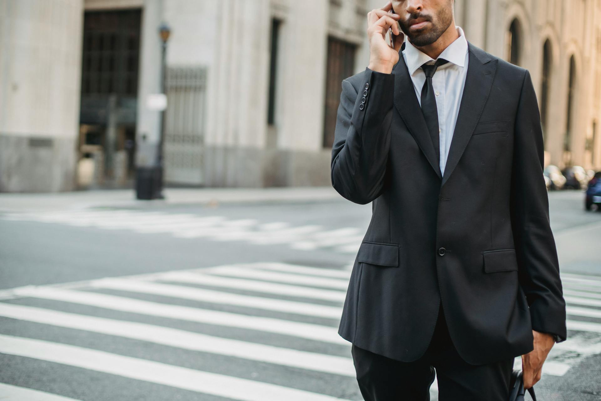 A businessman in a black suit talks on a phone while standing in a city street crosswalk.