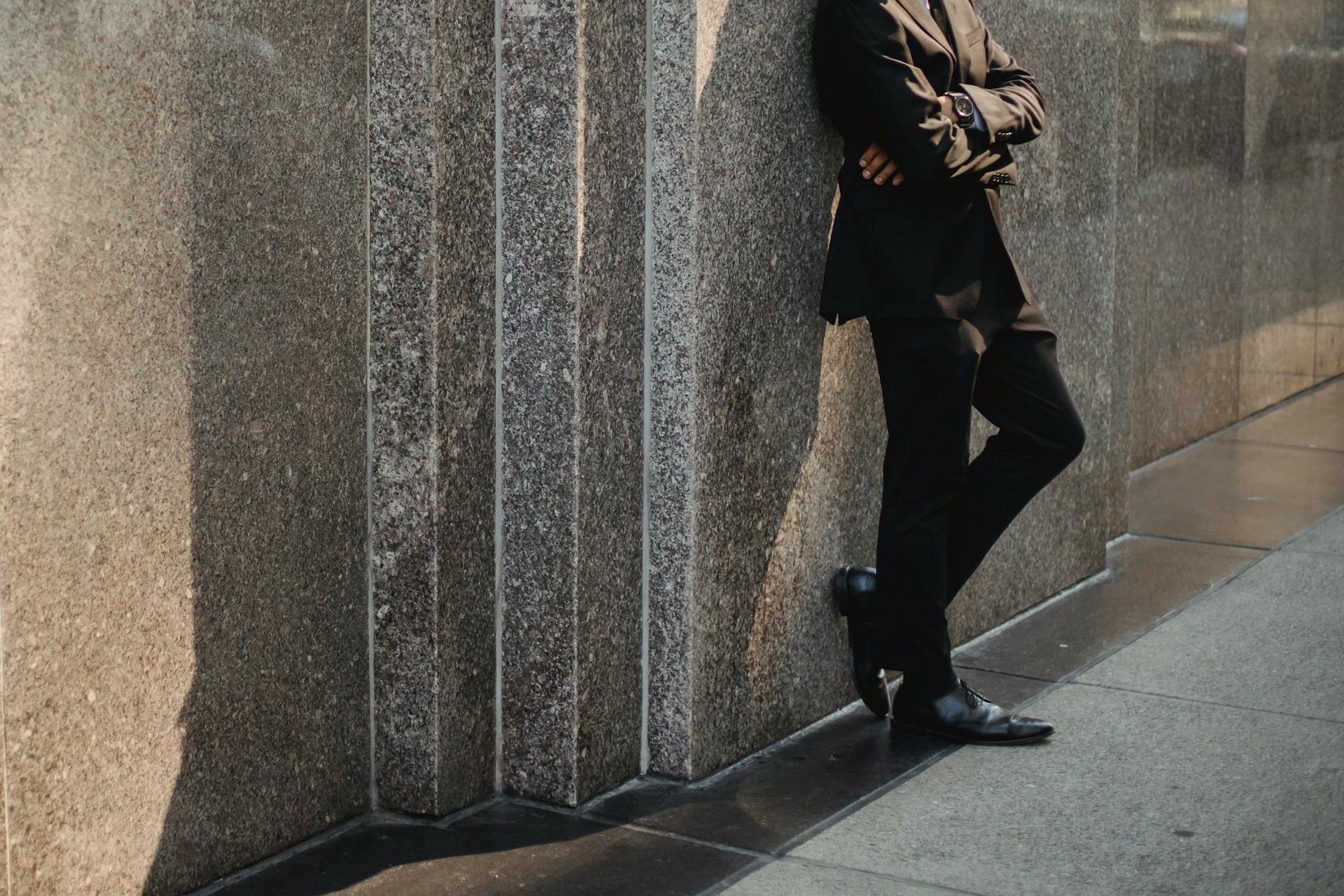 Elegant businessman in a suit leaning against a stone wall in an urban setting.