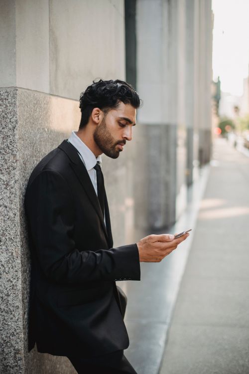 Free Businessman in a suit checking his phone while leaning against a building wall outdoors. Stock Photo