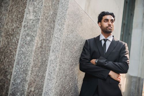 Free Businessman in a full suit with arms crossed leaning against a stone wall outdoors. Stock Photo