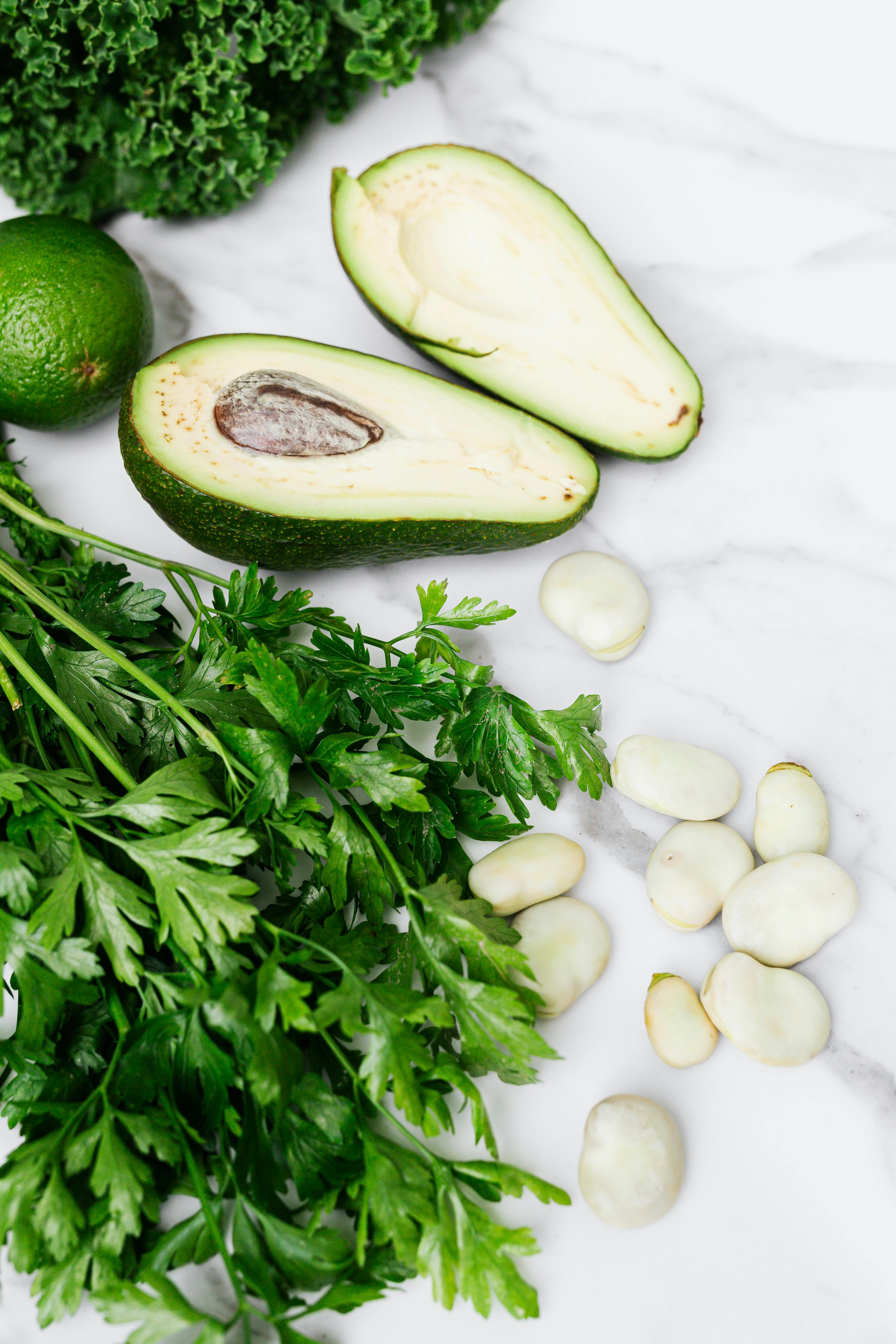 Crisp parsley, ripe avocado, broad beans, and lime on a white marble surface, a fresh culinary display.