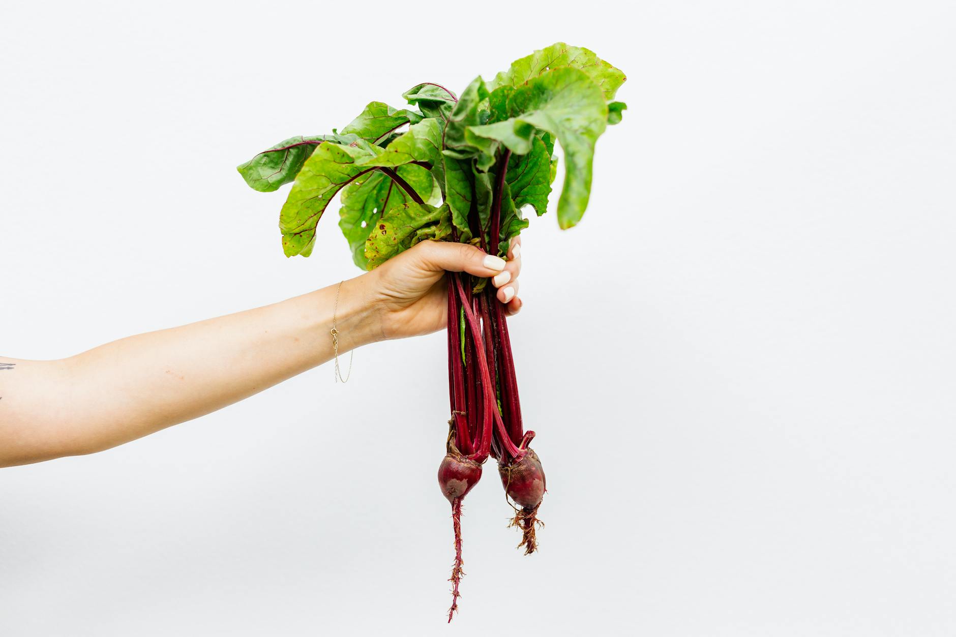 Hand holding vibrant fresh beetroots with green leaves against a simple white background.