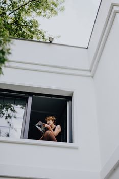 一名妇女坐在敞开的窗户旁，在户外现代风格的白色建筑中阅读杂志