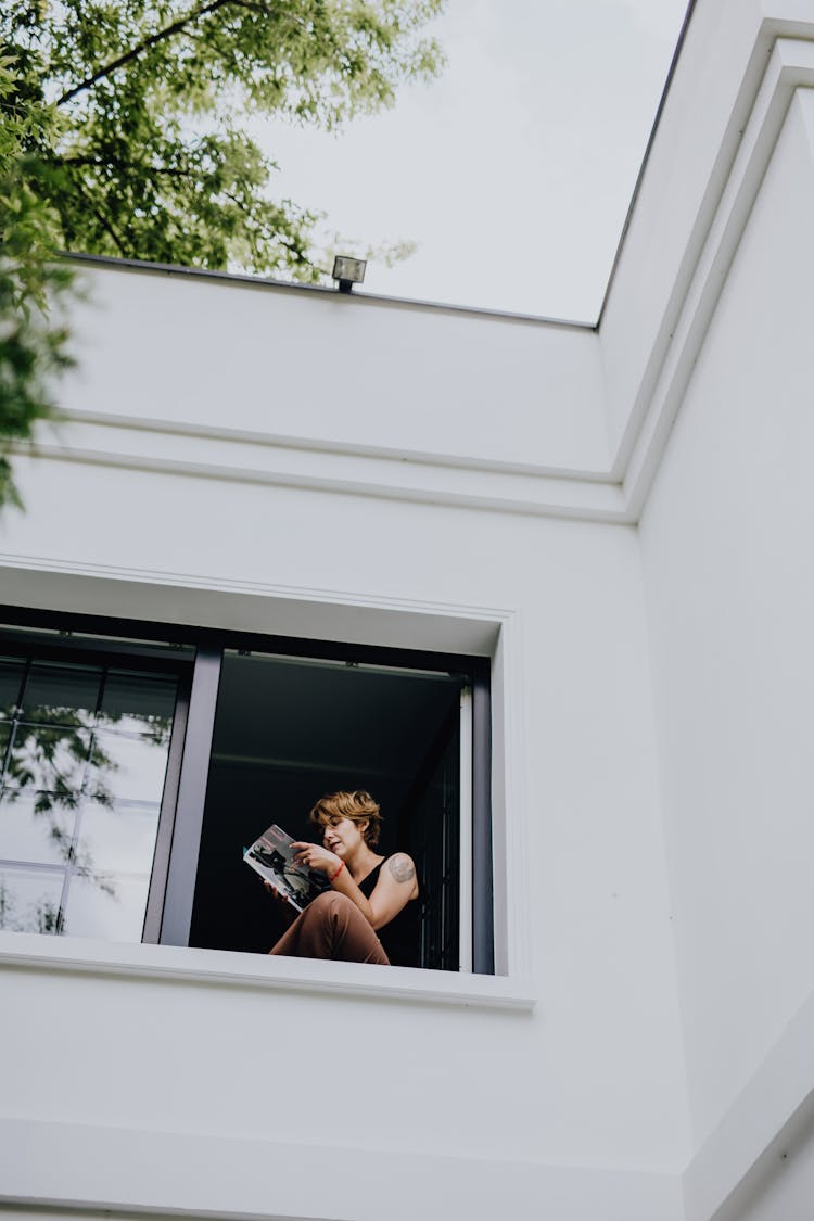 Girl Sitting In Open Window And Reading Magazine