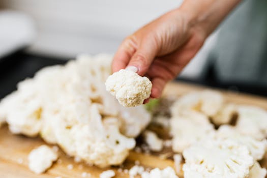 A hand holds a fresh cauliflower floret over a wooden board with scattered florets, showcasing a cooking preparation.