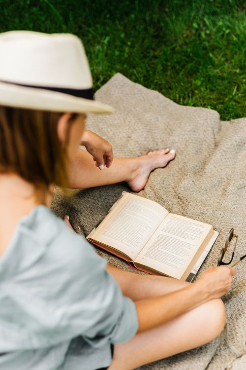 Free A woman in a hat enjoys reading a book on a blanket in the grass. Stock Photo