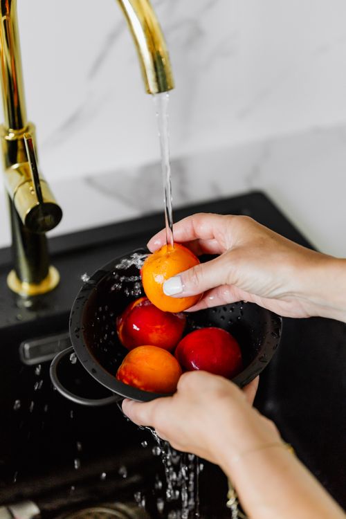 Free Hands washing fresh peaches under a brass tap in a modern kitchen setting. Stock Photo