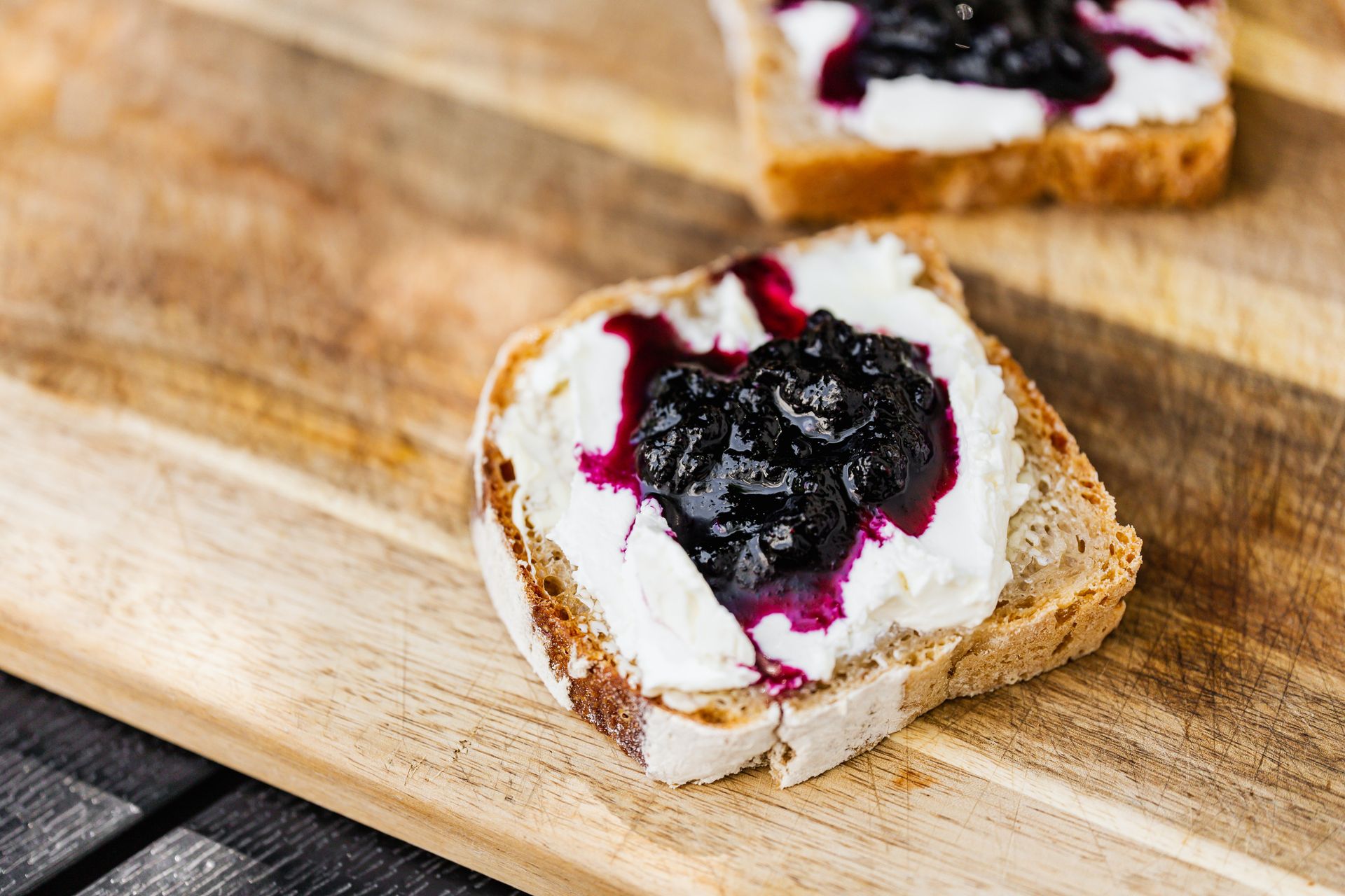 Delicious bread slice topped with creamy cheese and rich blueberry jam on a wooden cutting board.
