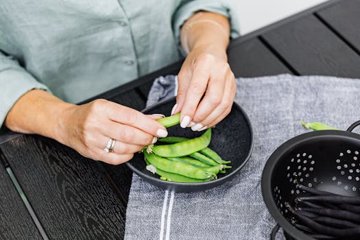 Hands peeling snap peas over a bowl, showcasing fresh ingredients.