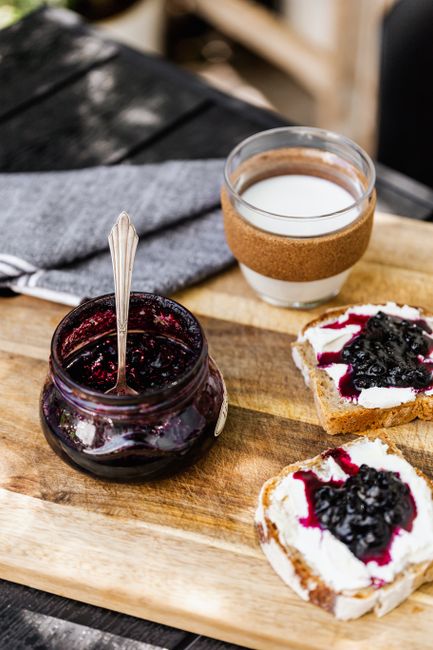 Two open jars of dark berry jam with visible crushed fruit pieces, one with a spoon resting in it and a slice of toast spread with jam beside them.