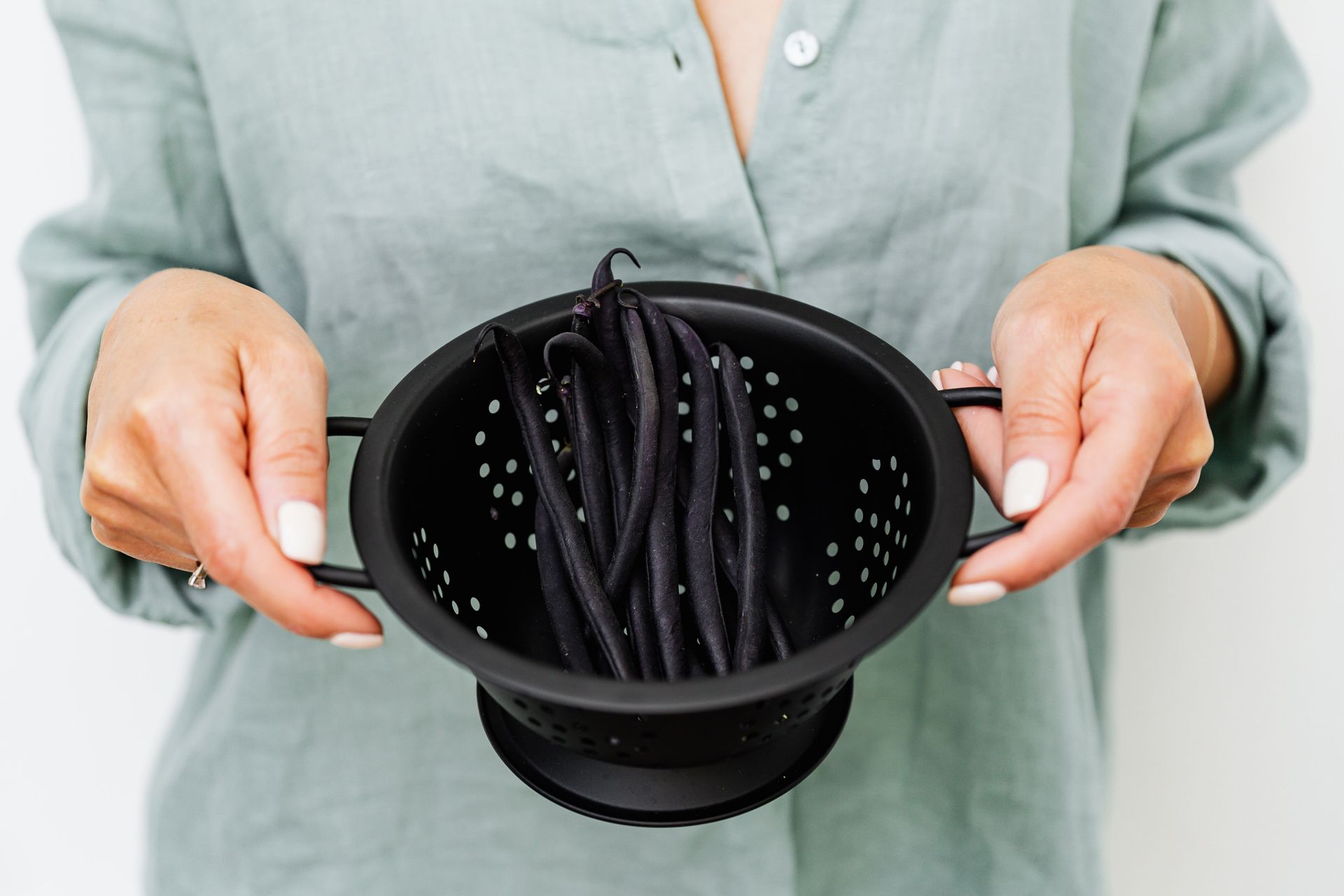 Woman holding a black colander with fresh black beans, emphasizing cooking and food preparation.