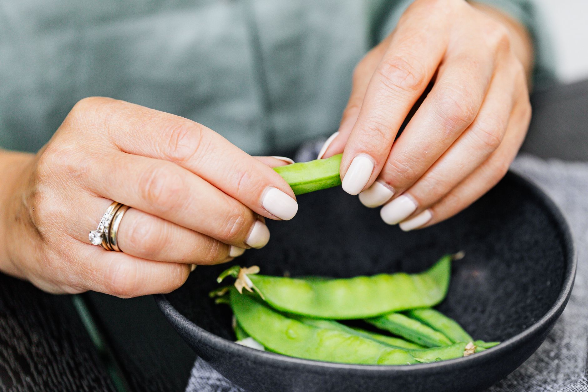 A woman with manicured nails shells fresh snap peas into a black bowl. - gut health reset