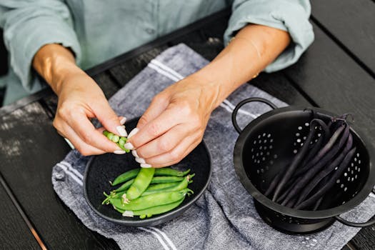 Close-up of hands shelling fresh snap peas over a black bowl outdoors.