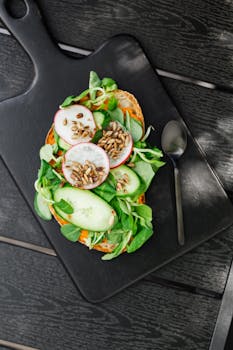 Healthy open-faced sandwich with radishes, cucumbers, greens, and seeds on a black platter.