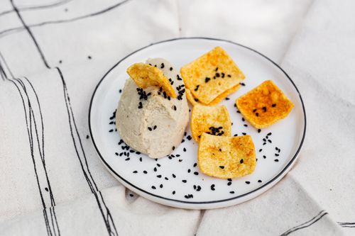 Free A plate of homemade hummus topped with black sesame seeds accompanied by crispy chips on a white tablecloth. Stock Photo