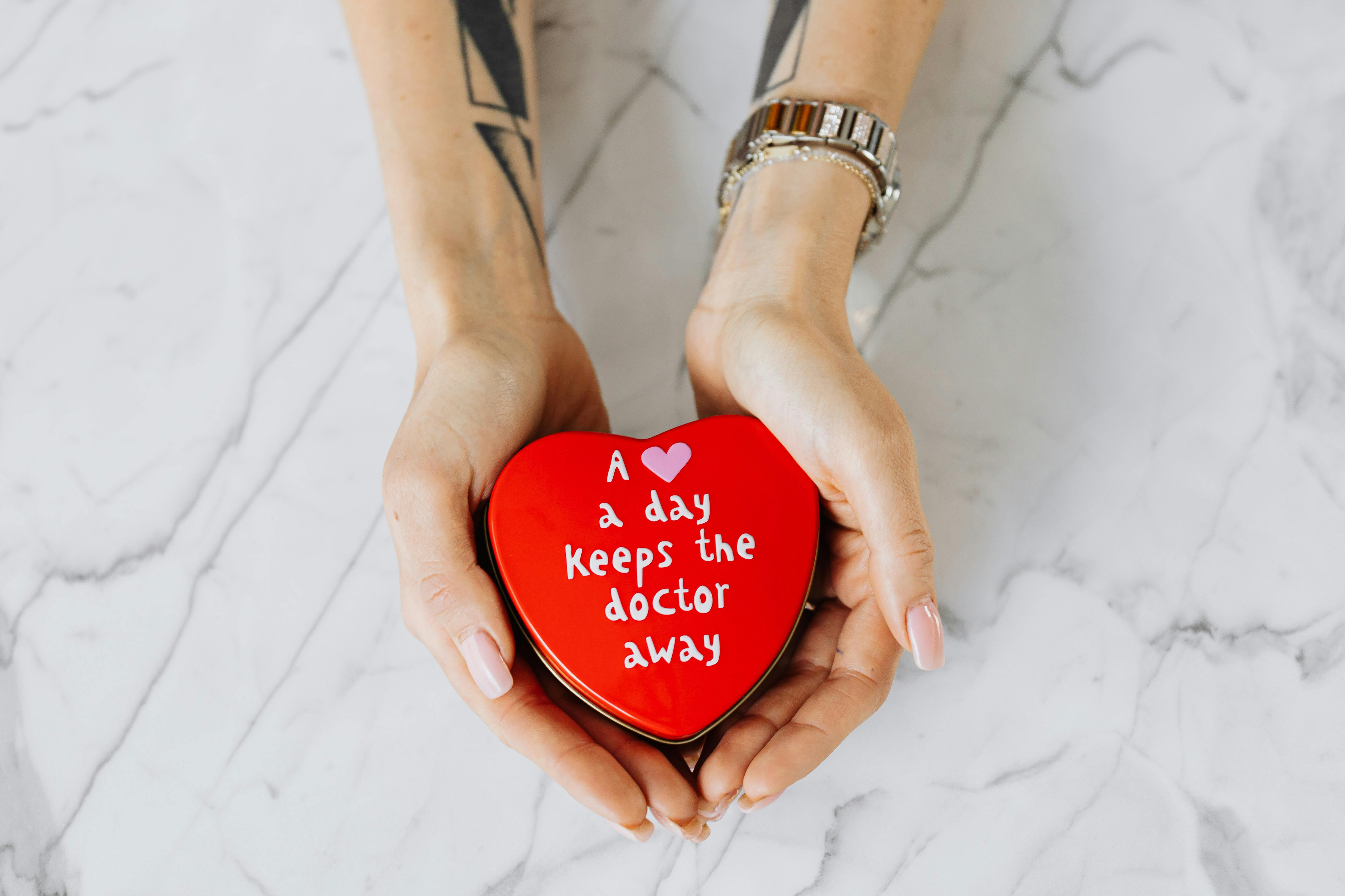 Hands holding a red heart-shaped tin with the message 'A heart a day keeps the doctor away' on a marble surface.