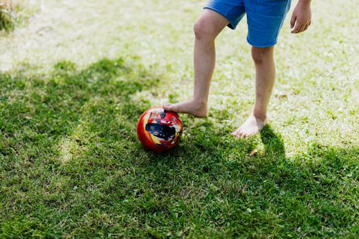A young child in blue shorts plays barefoot with a red soccer ball on green grass.