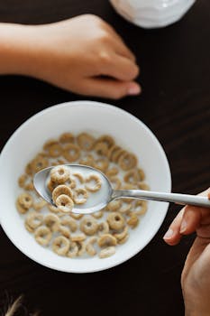 A spoon of cereal in milk, with hands and a bowl, captured indoors.