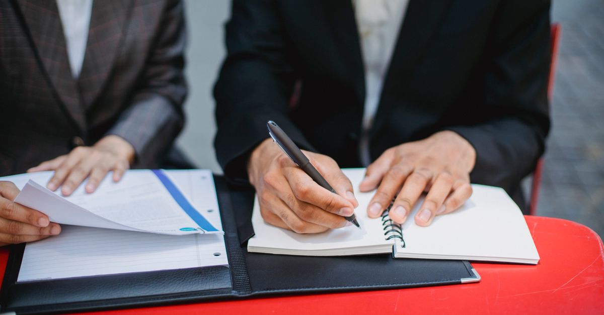 Two professionals in suits writing notes and reviewing documents at an outdoor meeting table.