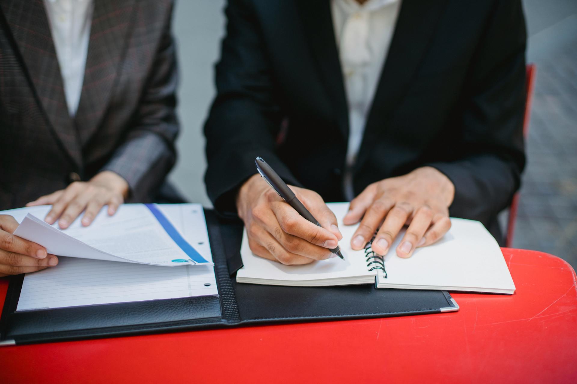 Two professionals in suits writing notes and reviewing documents at an outdoor meeting table.