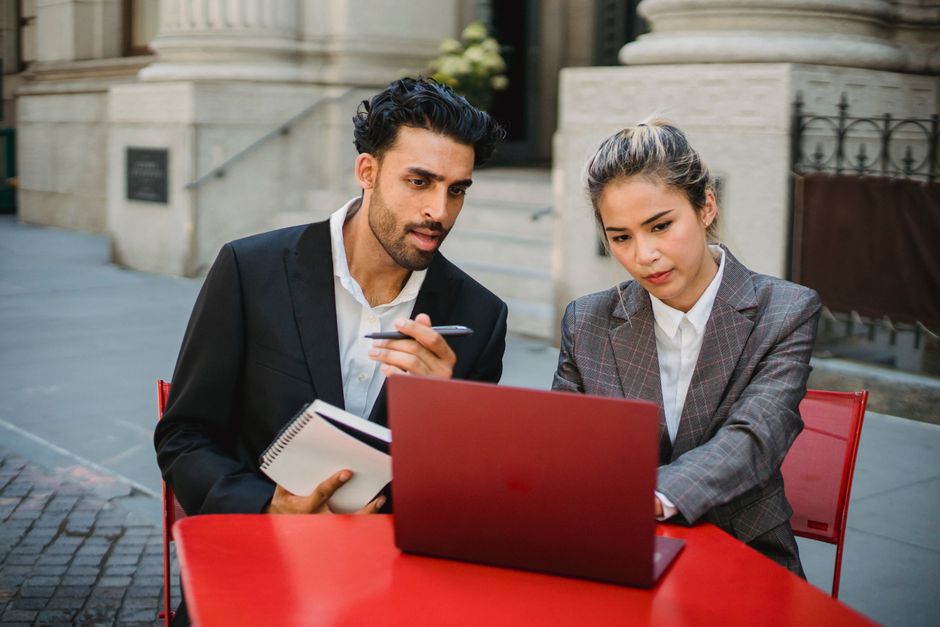Two professionals collaborate at an outdoor table with a laptop and notebook.