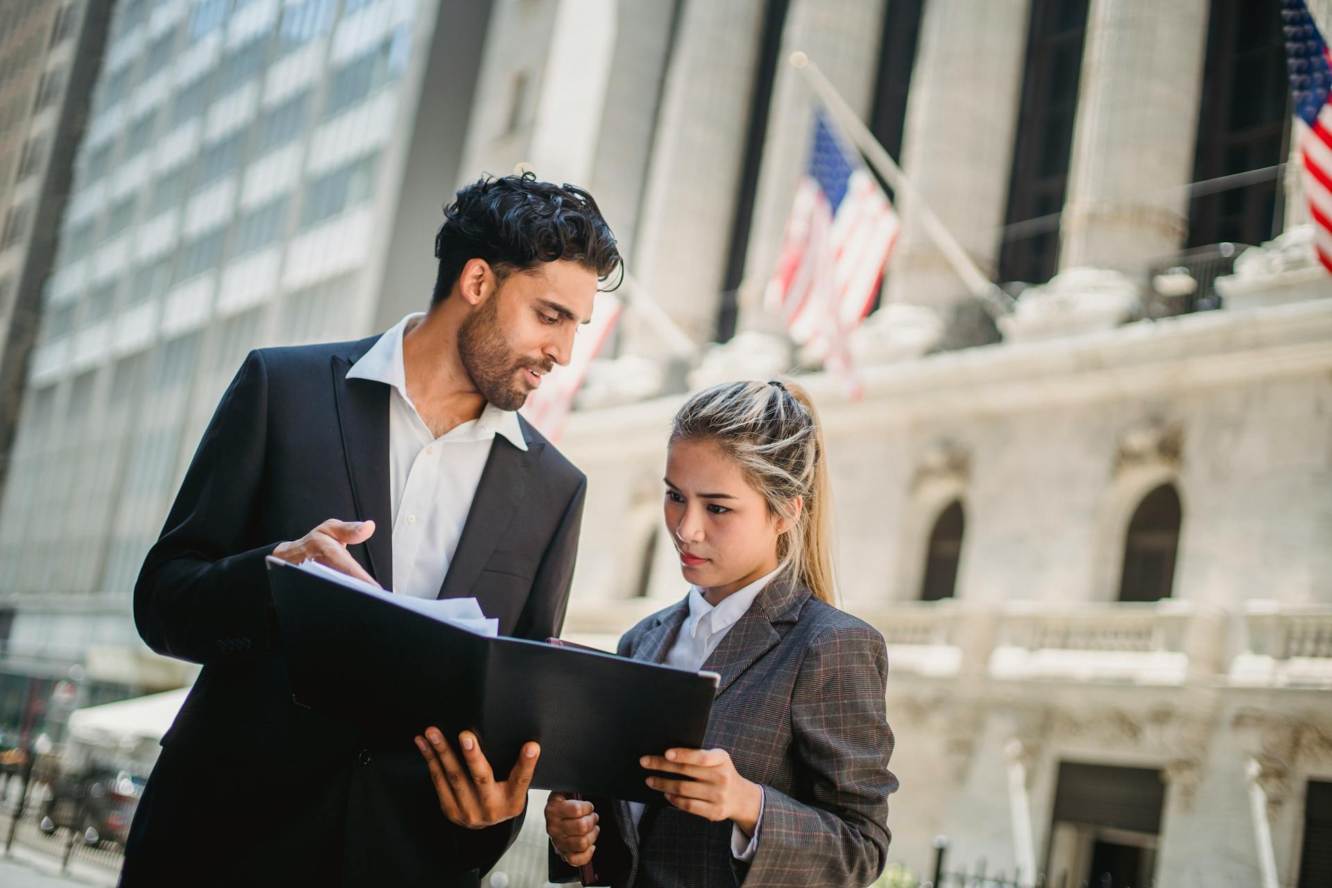 Two professionals in suits discussing documents outside a corporate building in daylight.