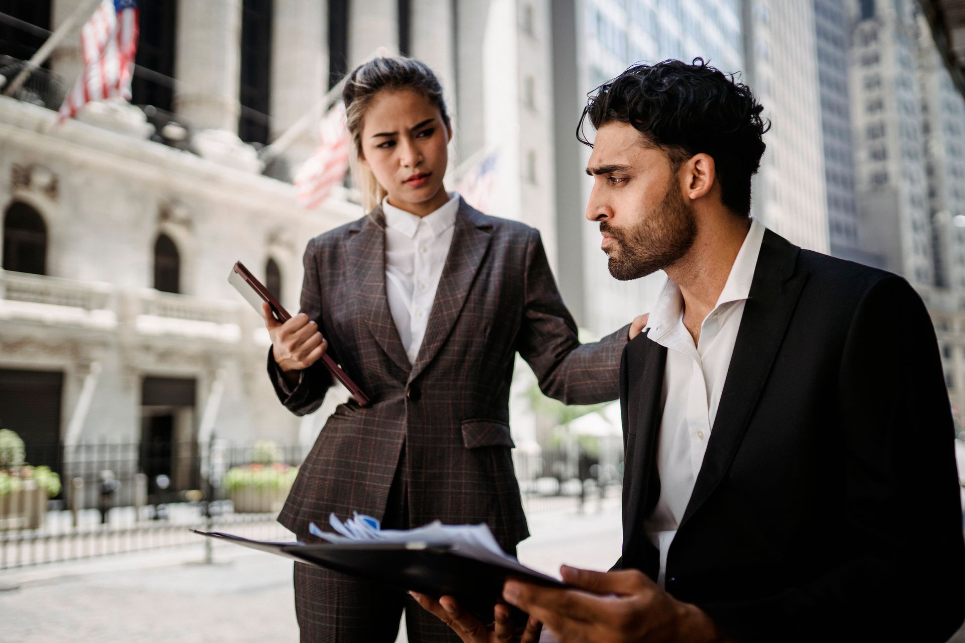 Two business professionals in formal attire discussing documents in a financial district setting outdoors.