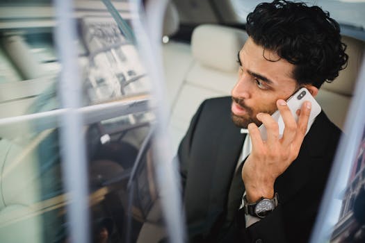 Man in black suit talking on a smartphone, sitting inside a car.