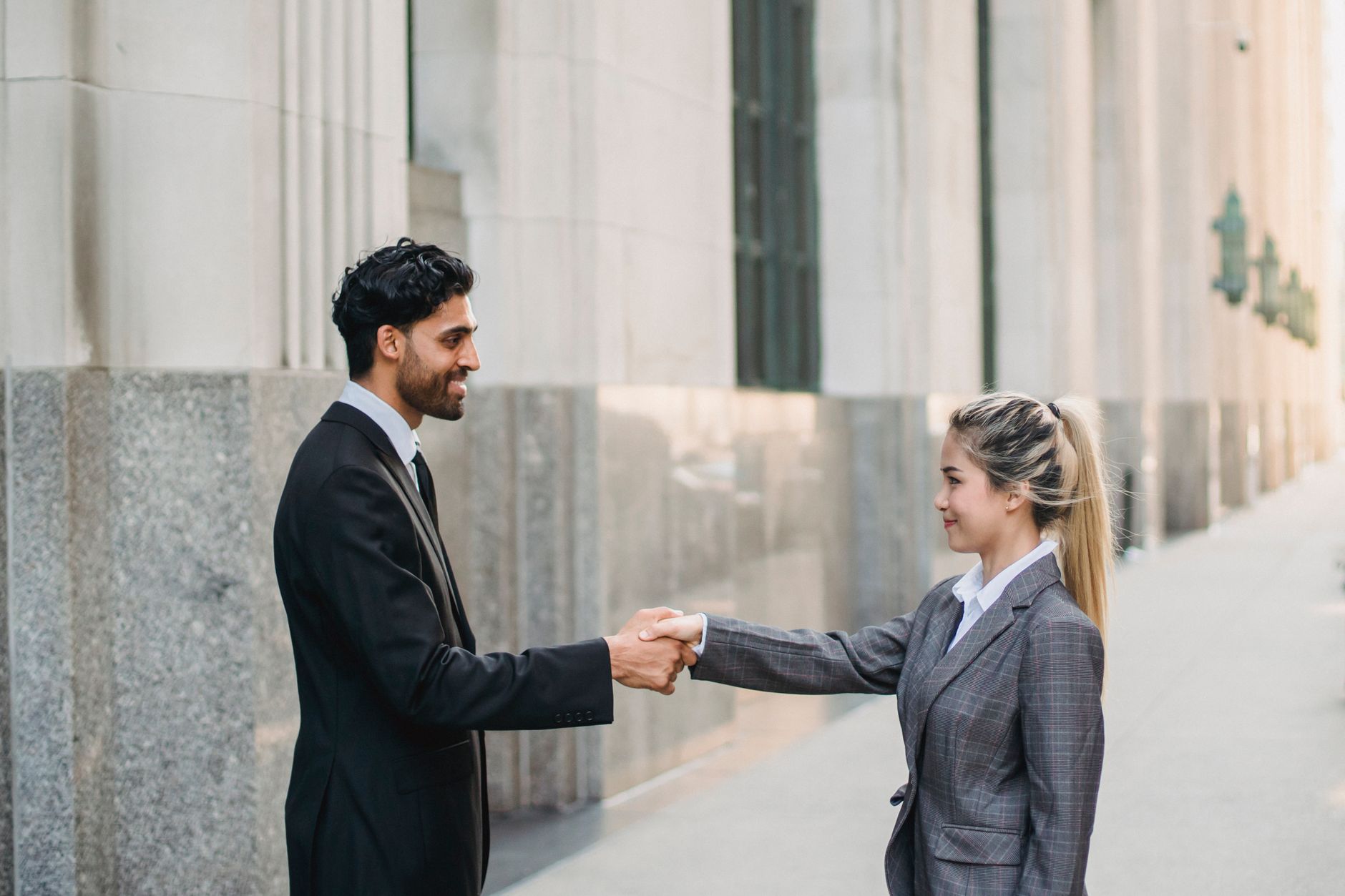 Two business professionals in suits exchanging a handshake outside a corporate building.